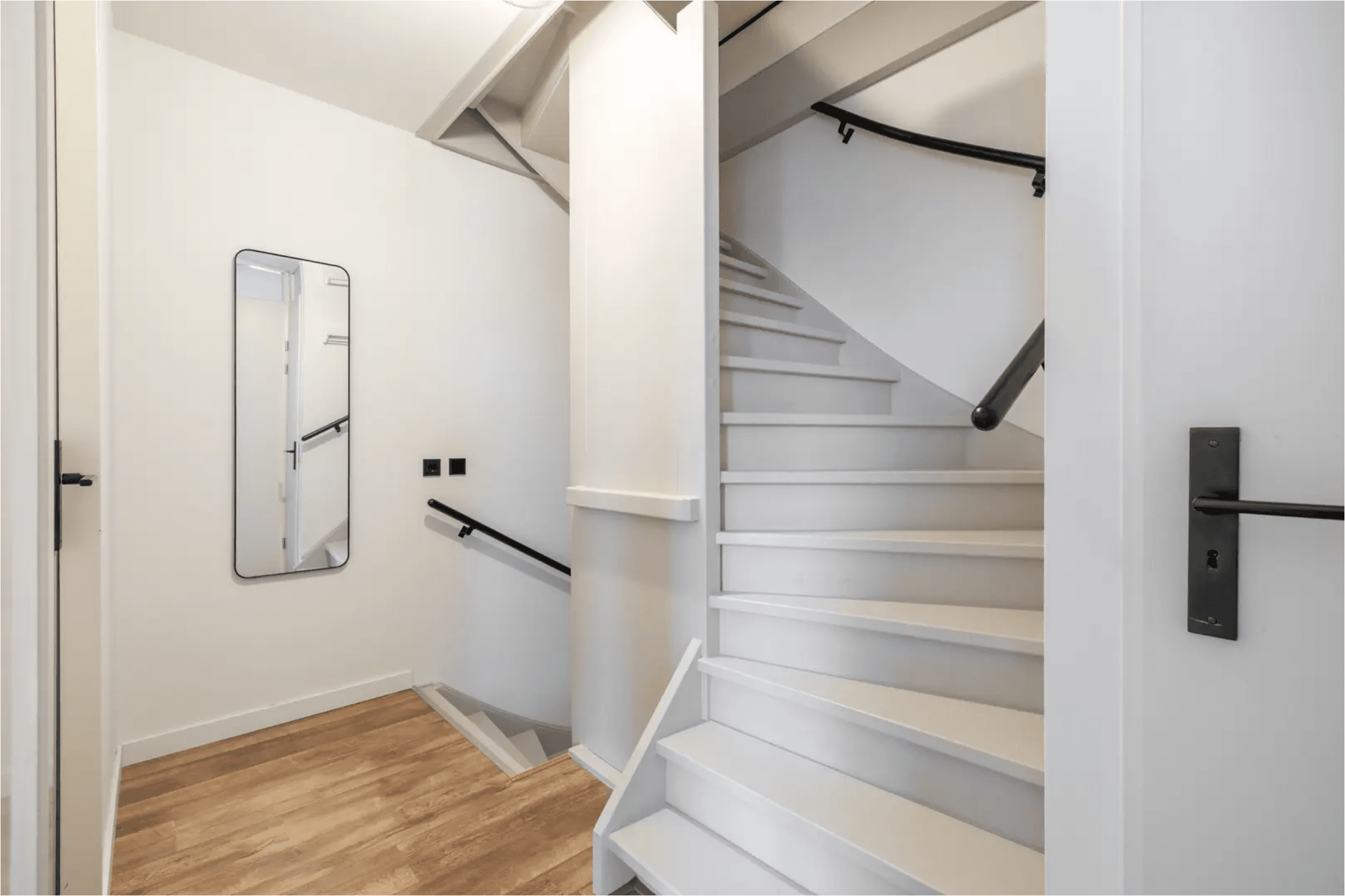 Minimalist white staircase with black handrails, wood flooring, and a tall wall mirror.