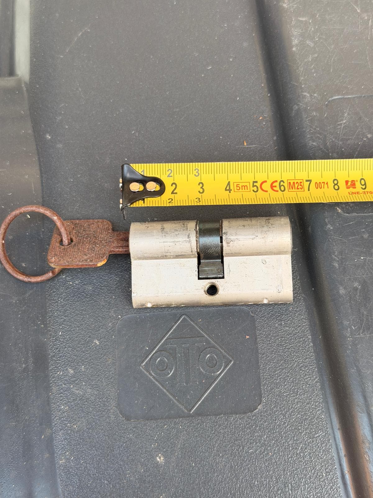 Metal door lock cylinder with a rusty key, measured by a yellow tape measure.