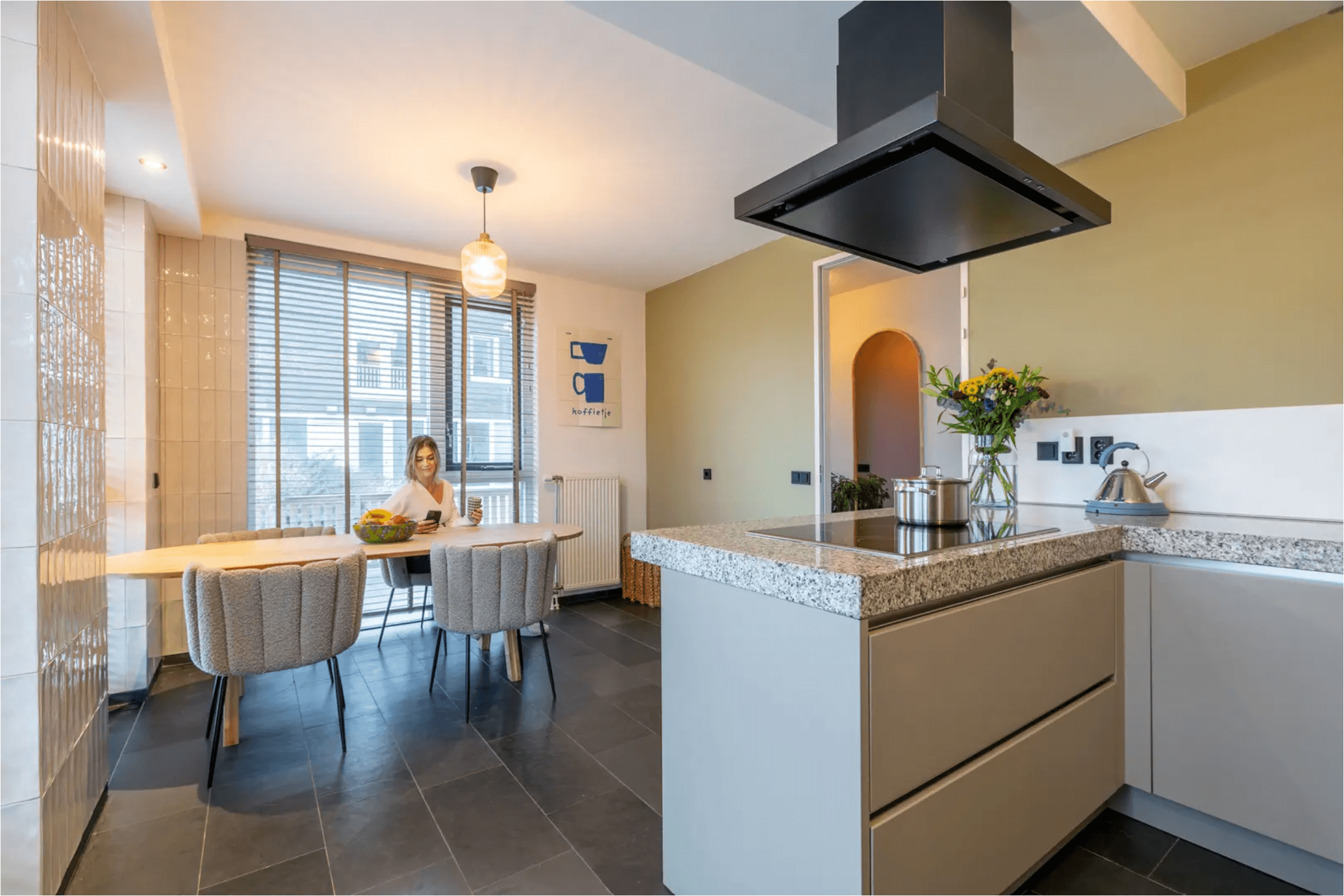 Modern kitchen with a granite island and a woman sitting at a sunlit dining table.