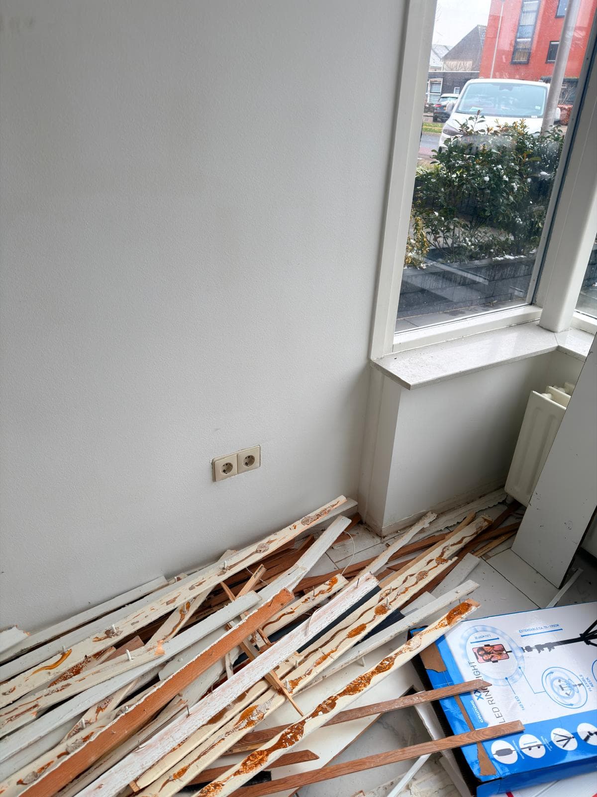 Discarded wooden baseboards with glue residue piled on a floor next to a white wall.