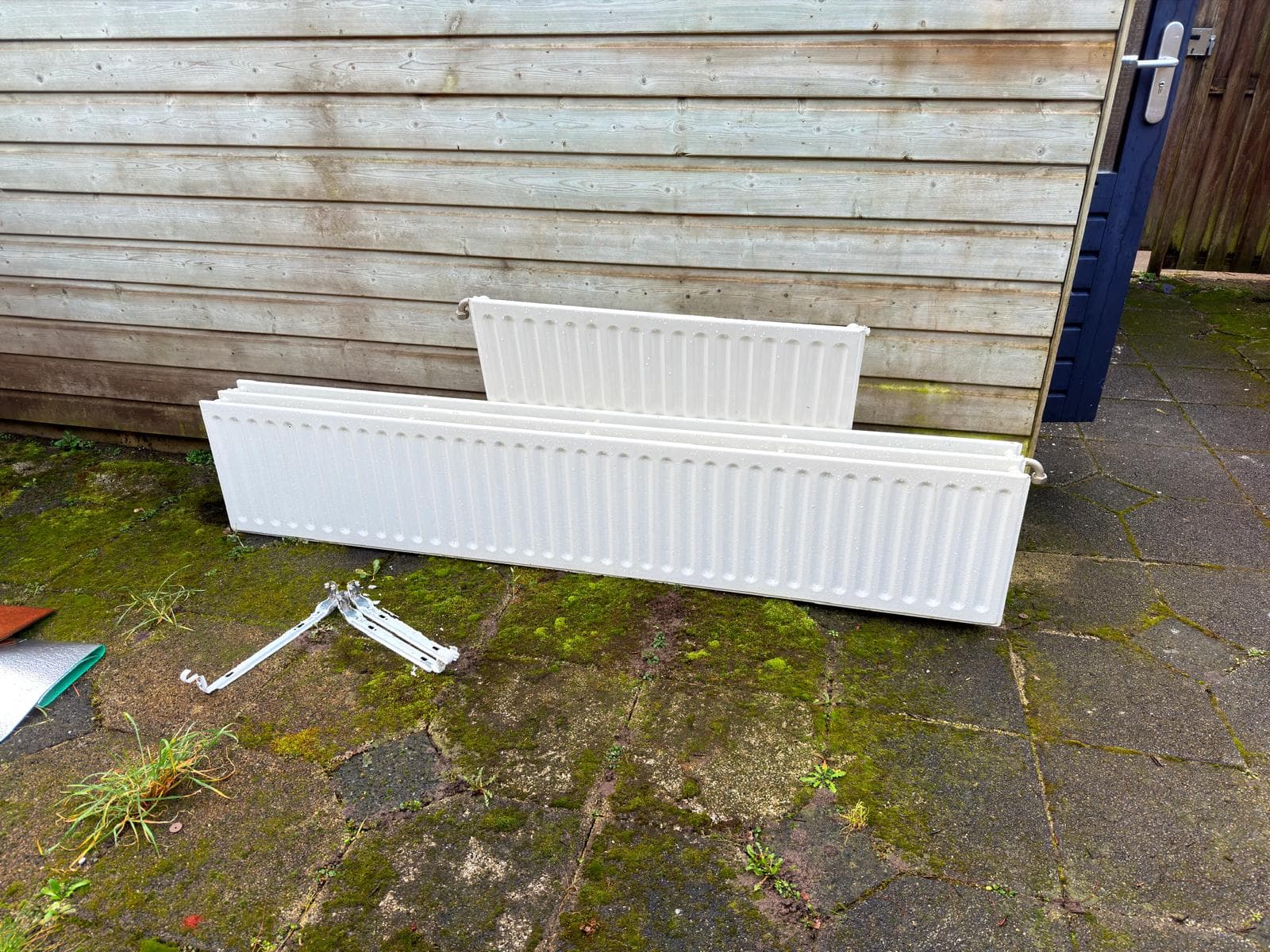 Three white radiators and metal brackets on a mossy patio against a wooden shed.
